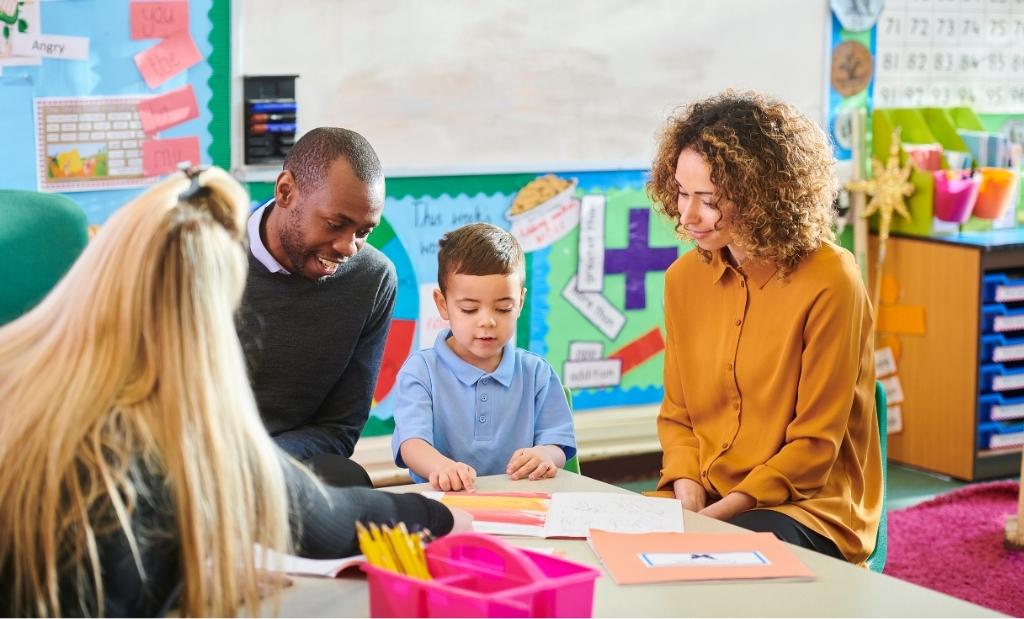 pais e professora sentados com uma criança na sala de aula durante uma reunião escolar.