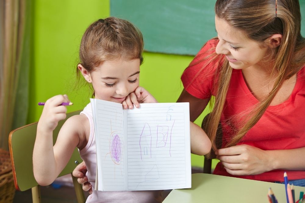 Menina mostra seu caderno de desenho para professora em sala de aula.