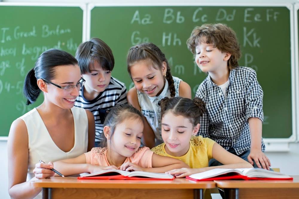 professora sorridente sentada com cinco crianças, todas olhando e lendo um livro aberto sobre a mesa, com quadro verde ao fundo.