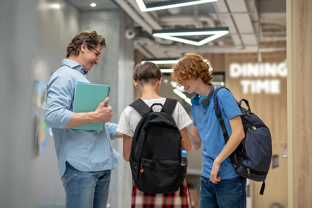 Professor segurando caderno enquanto conversa com dois alunos do ensino médio no corredor da escola.