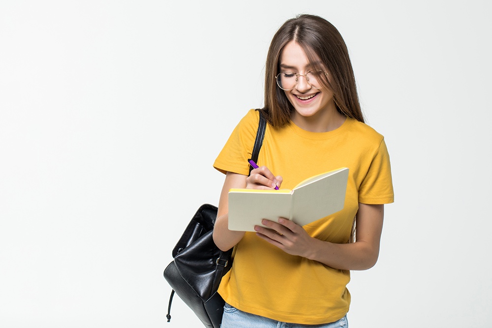 Menina jovem com mochila nas costas escrevendo em um caderno.