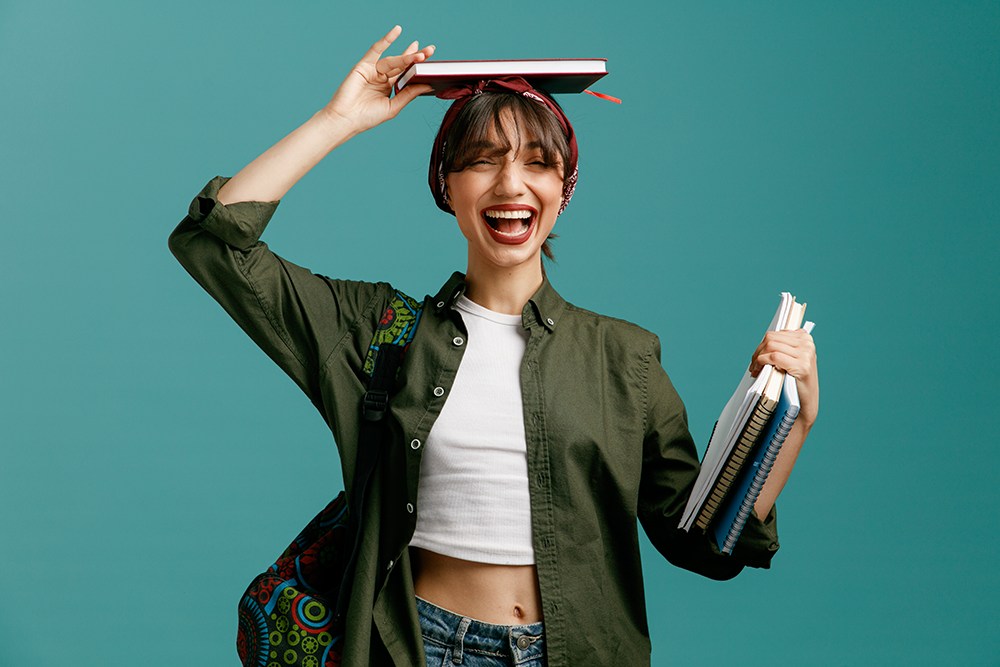 Mulher jovem segurando livros enquanto equilibra livro na cabeça.