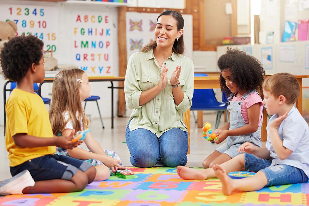 Professora de ensino infantil sentada em um tapete ao lado de quatro alunos conversando e brincando.