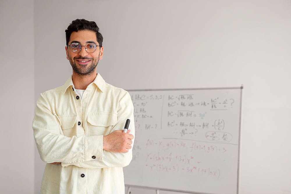 Professor de matemática de braços cruzados segurando caneta em frente a uma lousa.