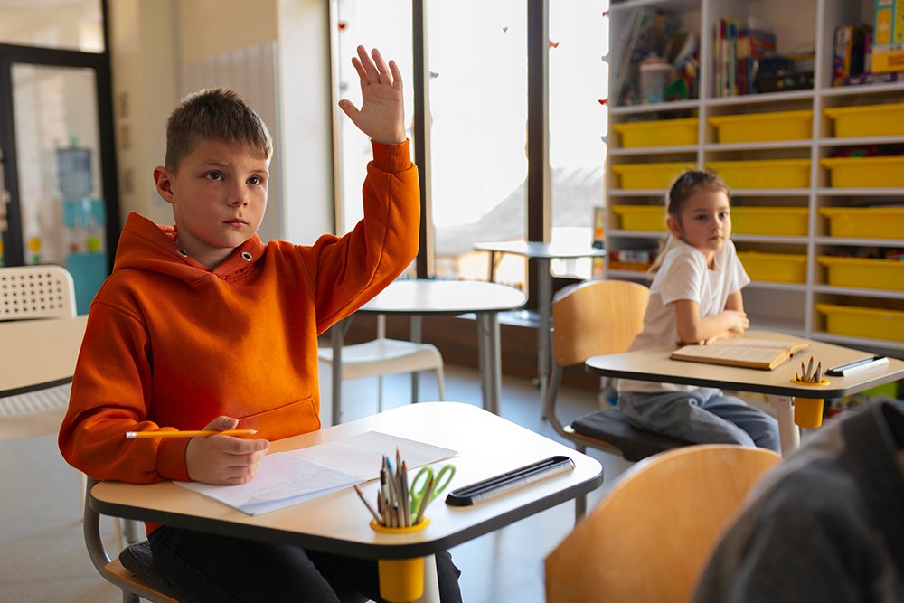 Aluno do ensino fundamental em sala de aula com a mão erguida para fazer perguntas.