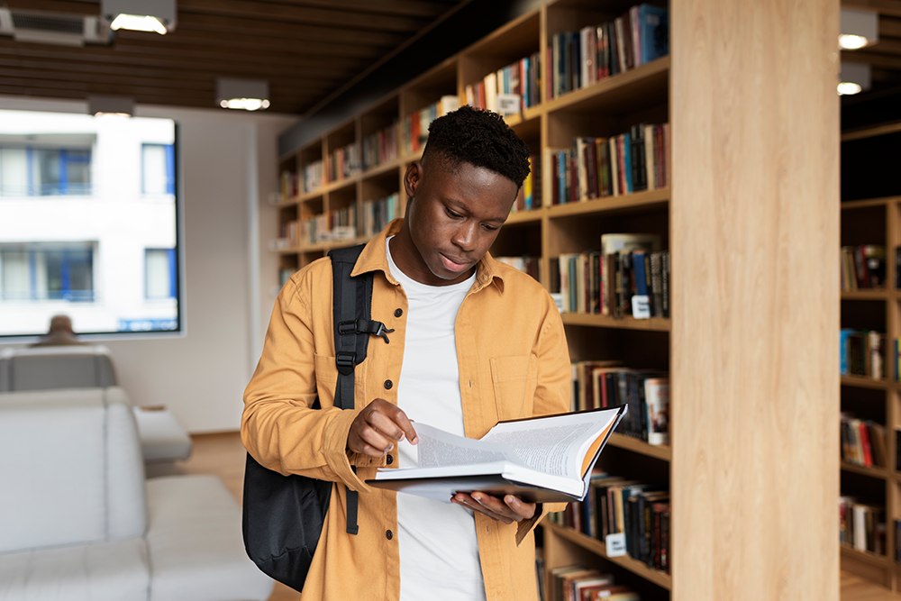 Estudante universitário com mochila nas costas folheando livro de capa dura em biblioteca.