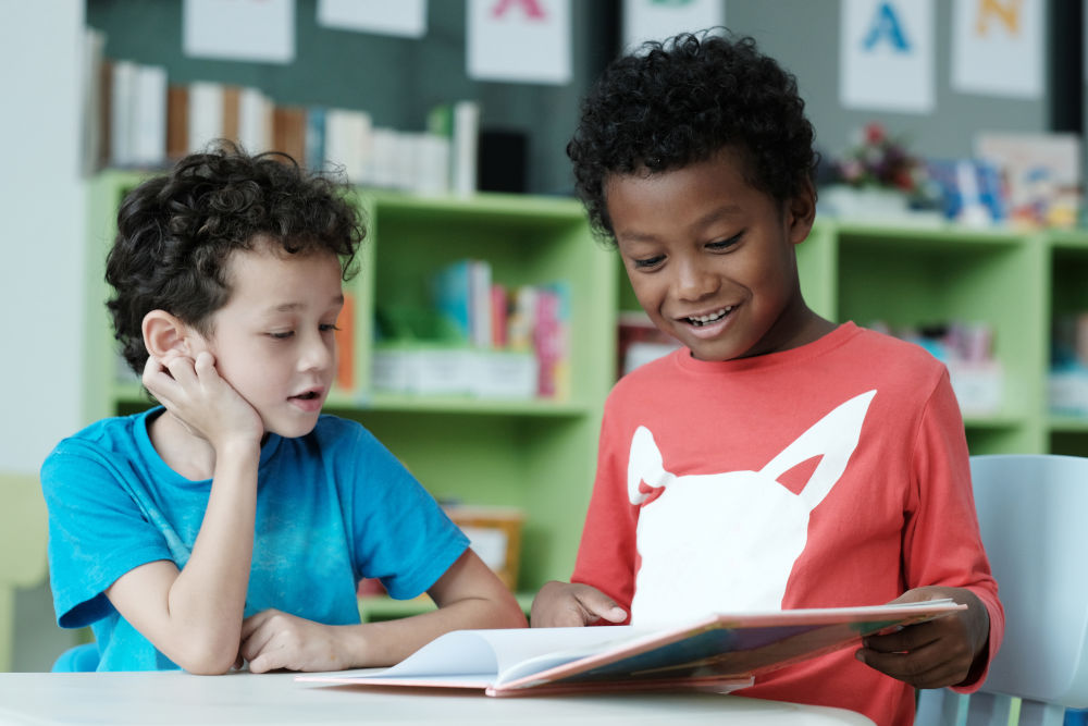 Dois meninos sentados em uma mesa escolar lendo um livro didático