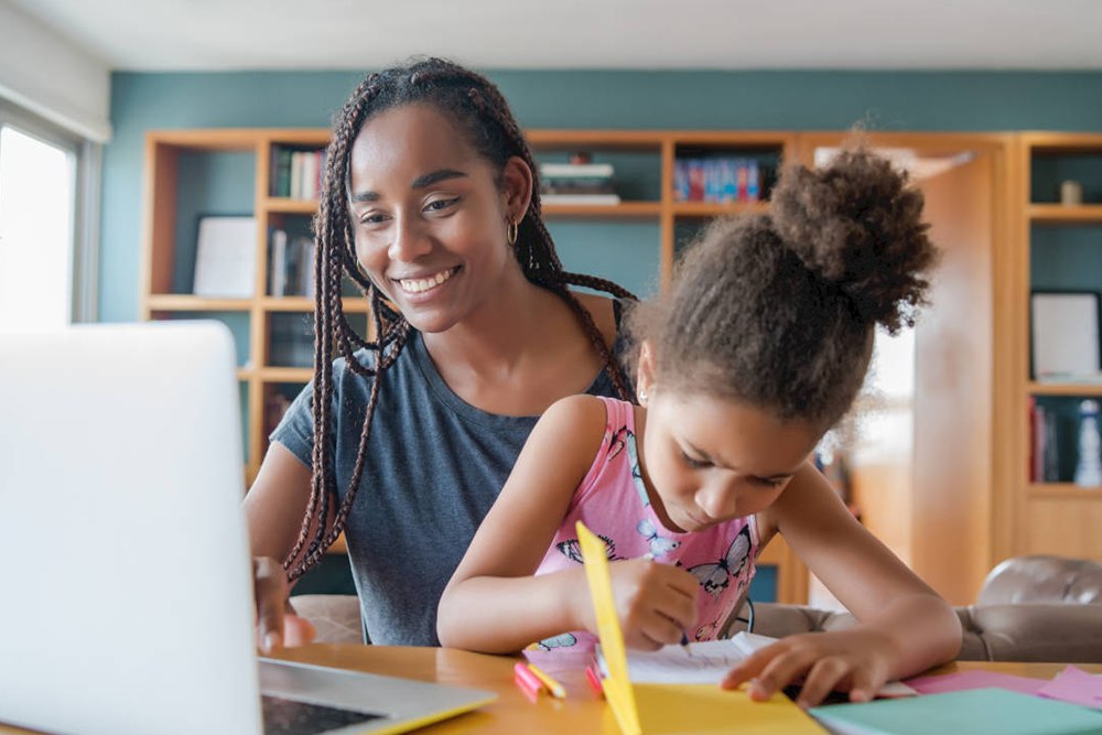 Menina sentada no colo da mãe enquanto desenha em um caderno e a mãe usa o computador.