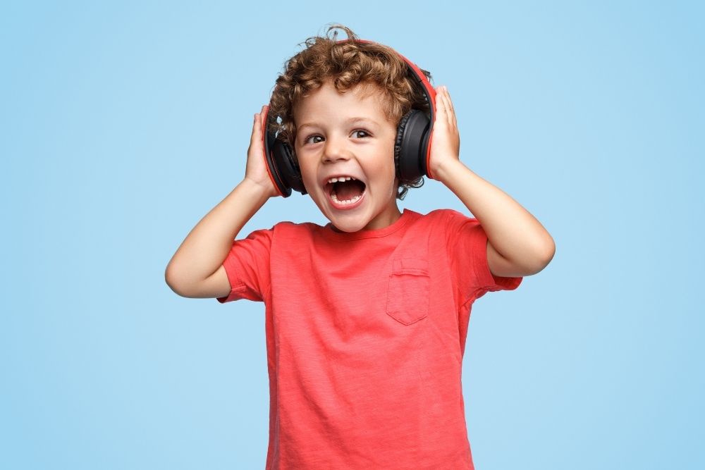 Menino com a boca aberta, vestindo uma camiseta vermelha, com as mãos em um fone de ouvido, sob um fundo azul.