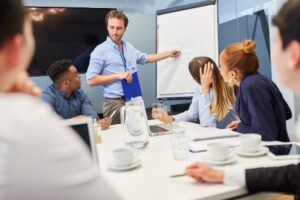 Homem de pé em frente a uma equipe, realizando uma apresentação em um quadro branco em um ambiente de escritório. Ao redor da mesa estão várias pessoas sentadas, prestando atenção, com laptops e xícaras.